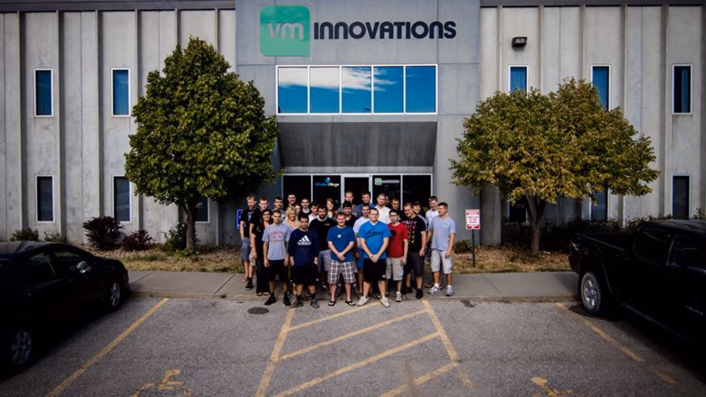 Group of about 25 people standing outside VMI Innovations building entrance between two trees.